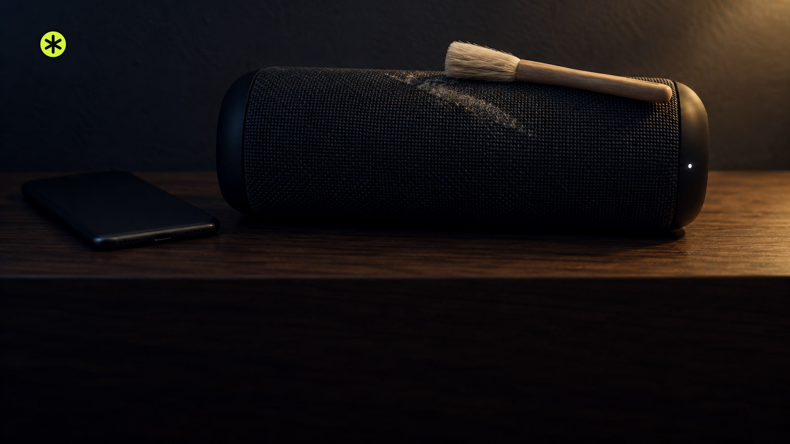 Charcoal fabric Bluetooth speaker on a walnut console with a soft brush resting on its grille and a phone face down beside it, lit by a low warm lamp.