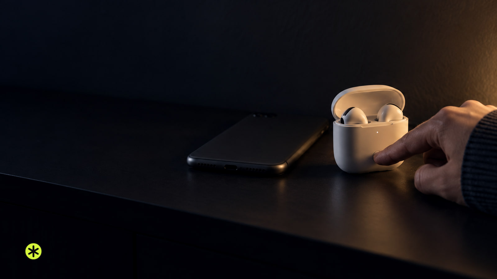 White earbuds in an open case on a black console at night, a fingertip pressing the button on its back beside a face down phone, warm lamp lit.