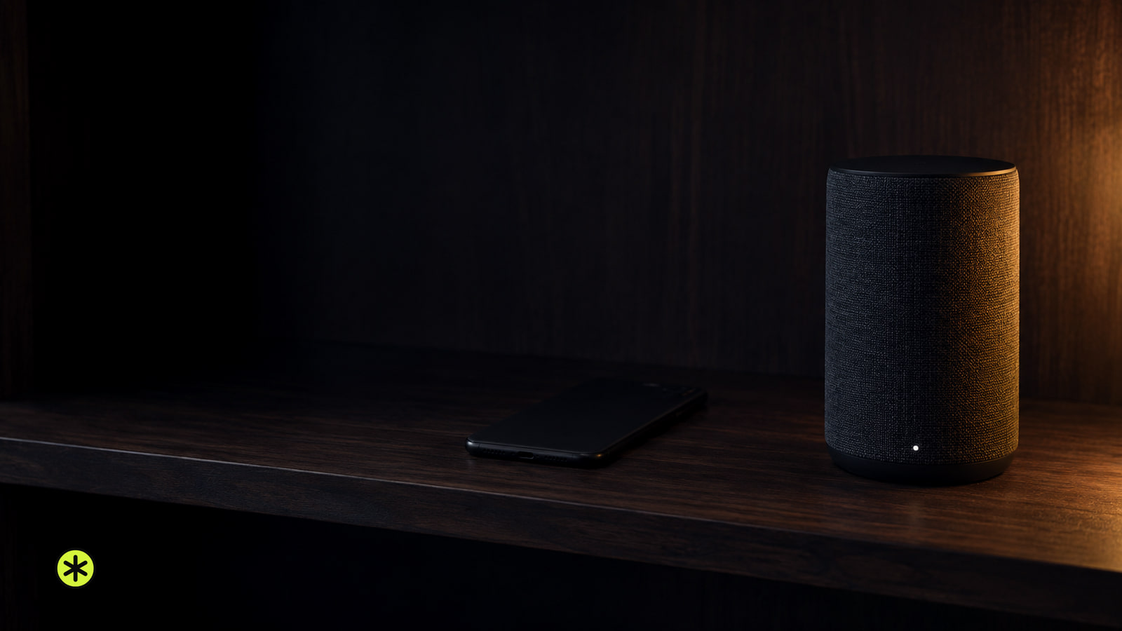 Charcoal fabric wireless speaker with a small white light on a walnut bookshelf, a phone face down nearby, lit by one low warm lamp at night.