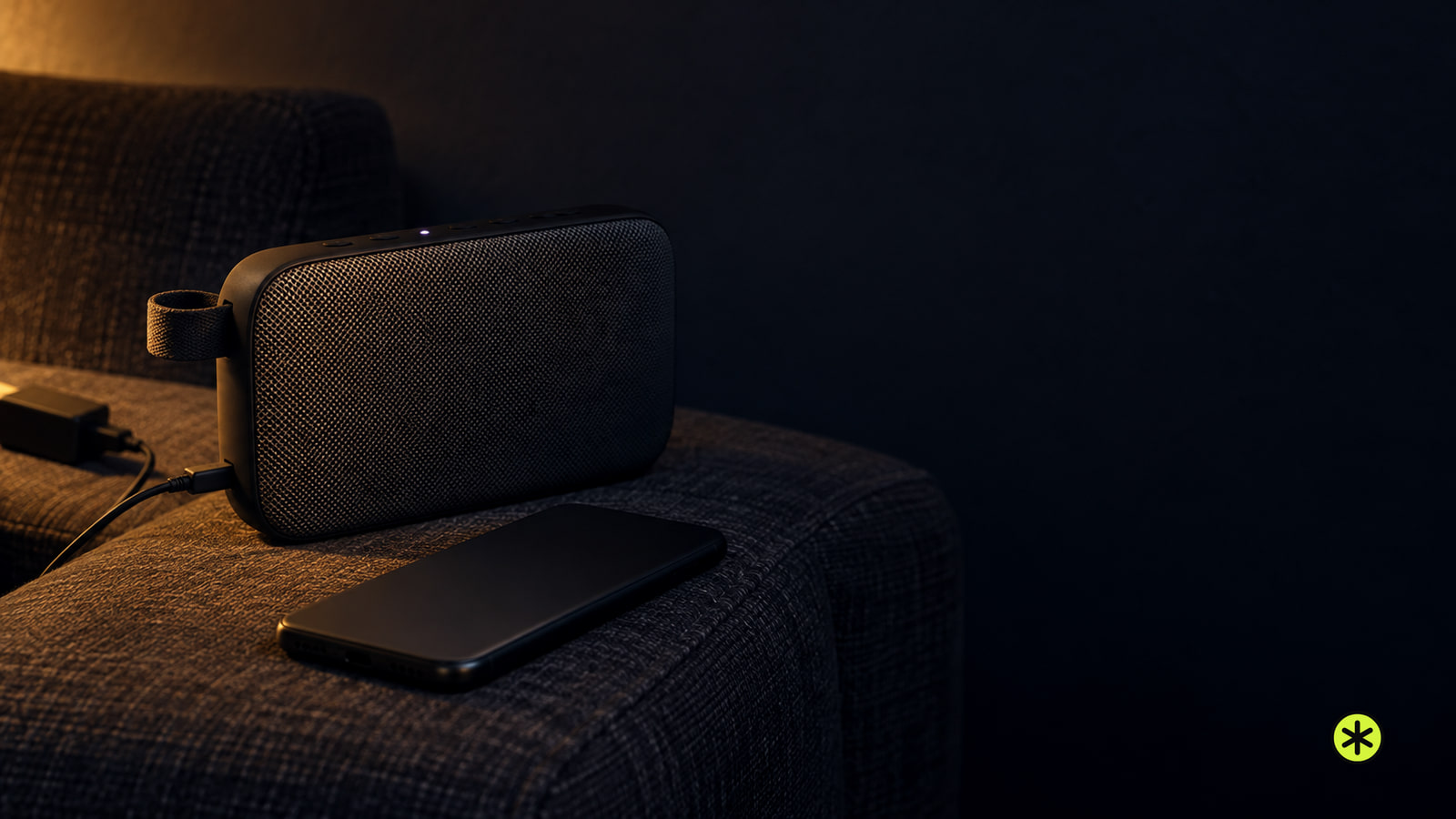 Charcoal portable speaker with a small white light beside a face-down phone on a dark armchair arm, lit by one warm lamp at night.