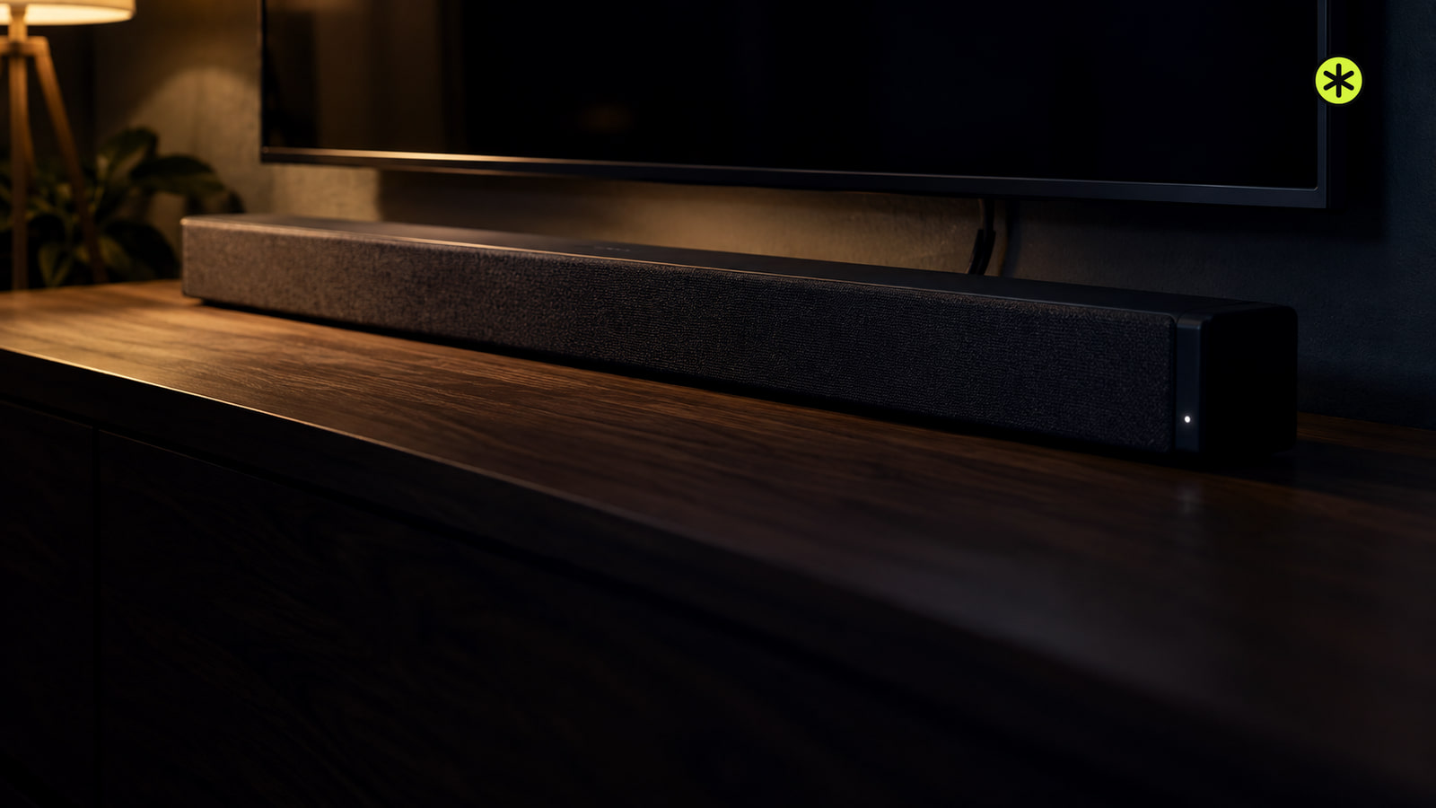 Charcoal fabric soundbar on a walnut console below a dark television at night, with a warm lamp lighting it from the upper left.