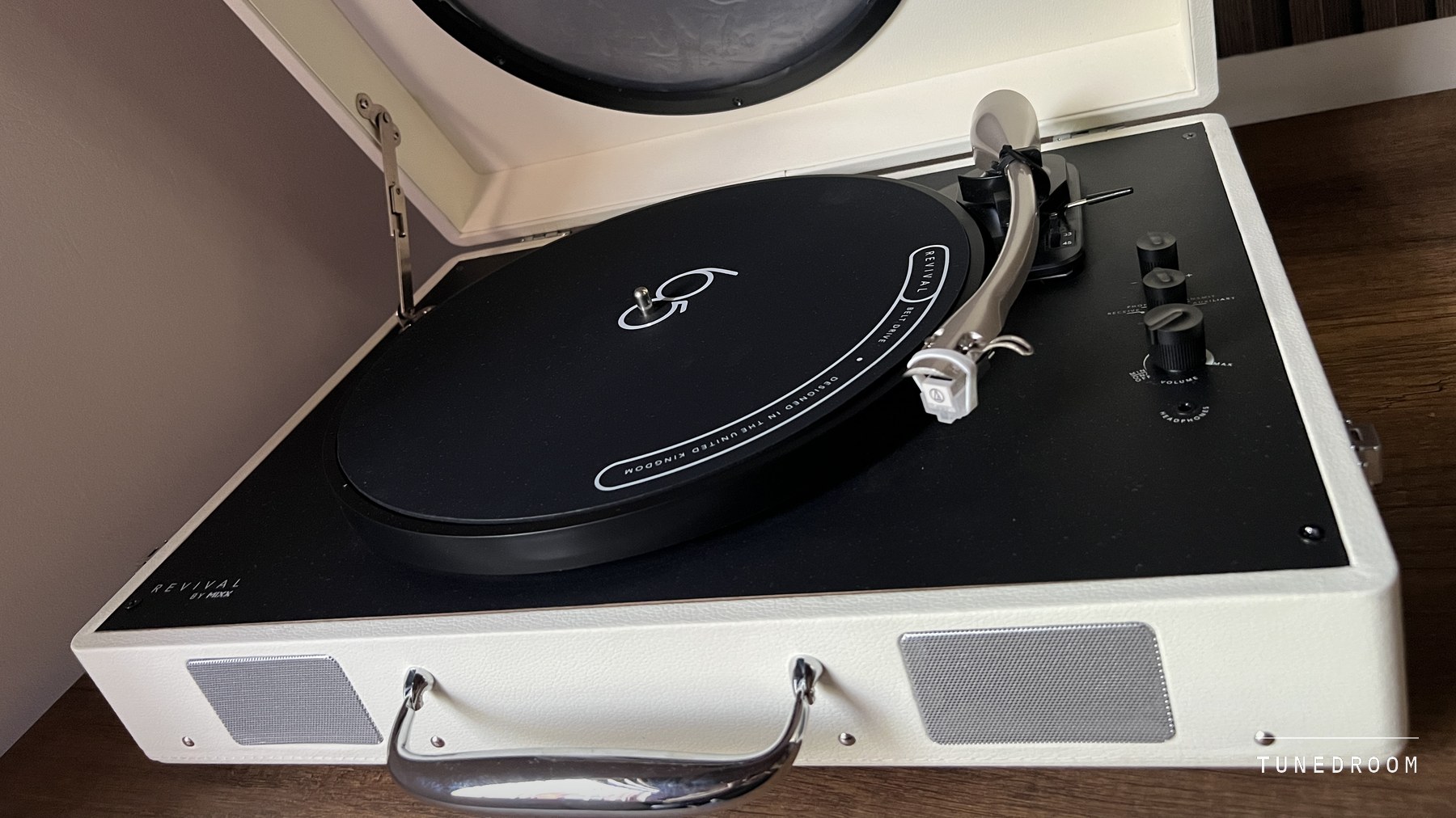 The cream MIXX Revival 65 open on a wooden sideboard, lid up, with the S-shaped tonearm and control knobs on the black deck and a speaker grille either side of the chrome handle