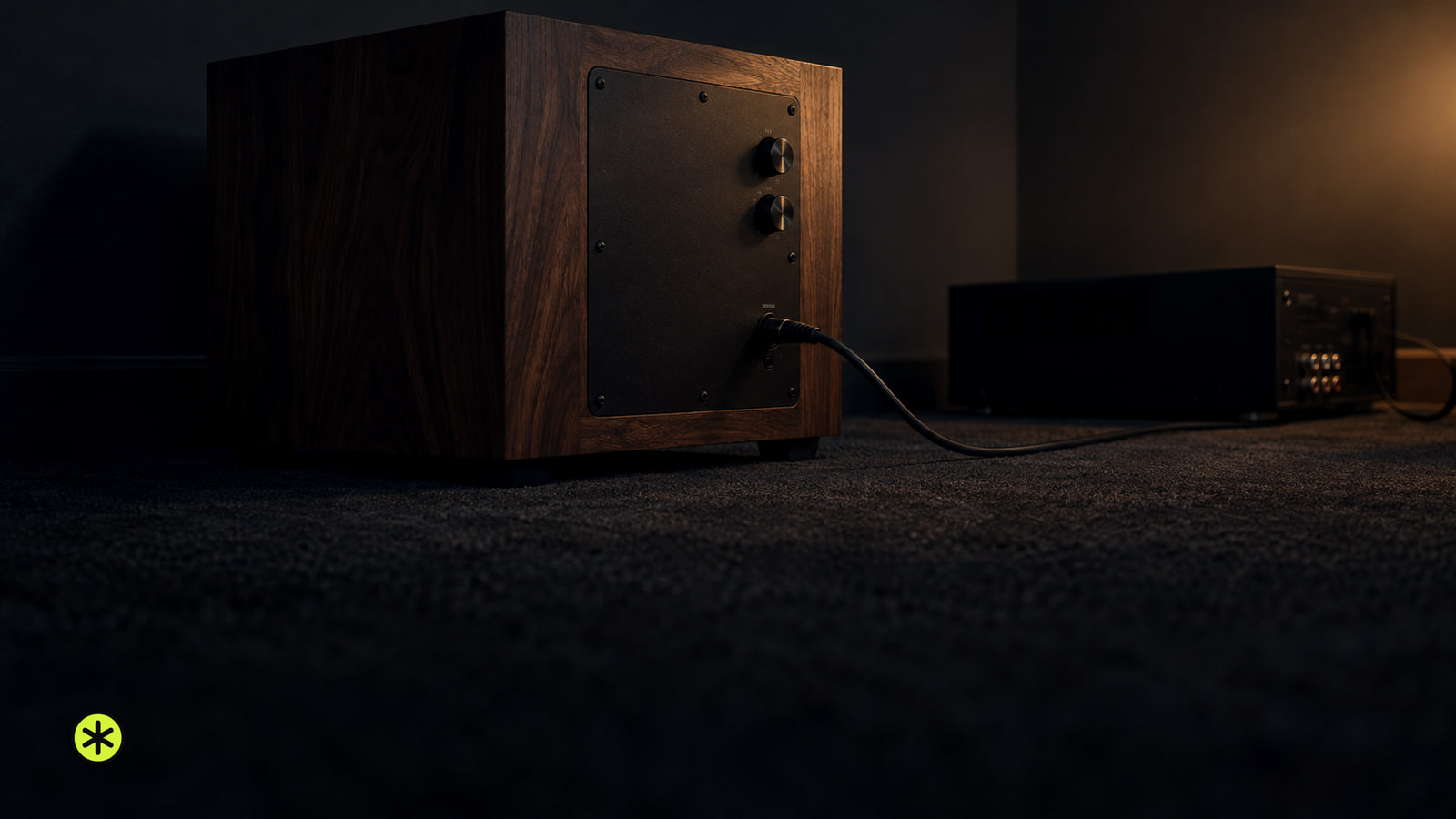 Walnut subwoofer on a charcoal rug seen from behind, two round knobs on its black rear plate and a cable to a receiver, lit by one warm lamp.