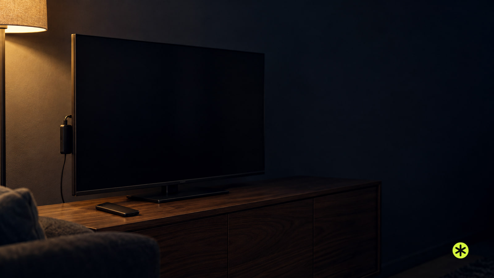 Dark flat television on a walnut sideboard with a small black streaming stick at its side and a phone face down, lit by one warm lamp at night.