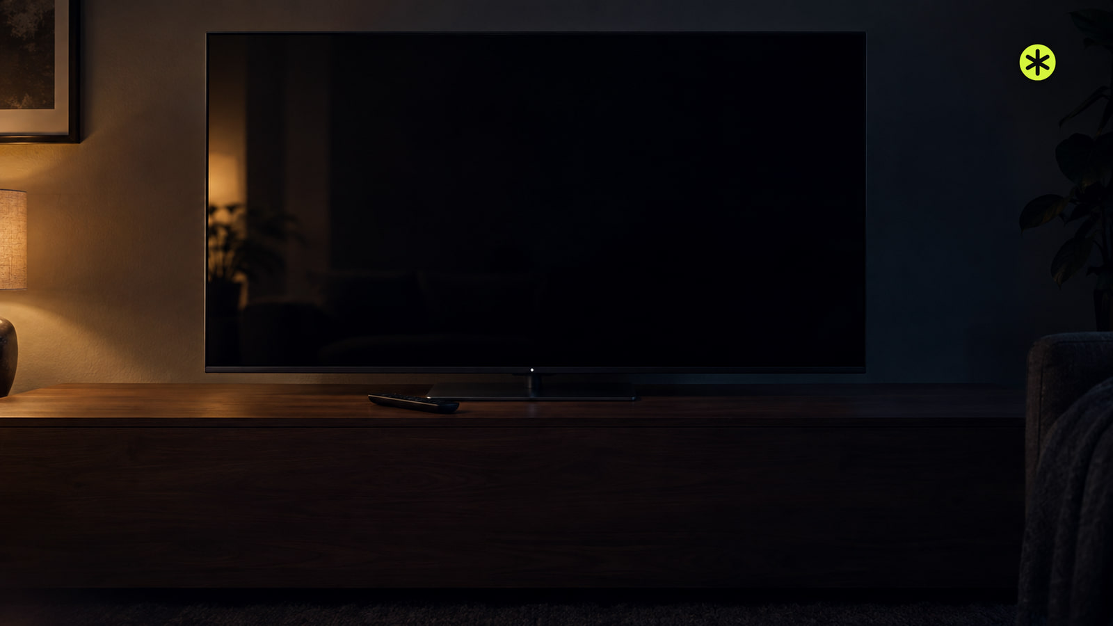 Black television screen on a walnut console at night with a slim remote in front, a warm lamp lighting it from the left.