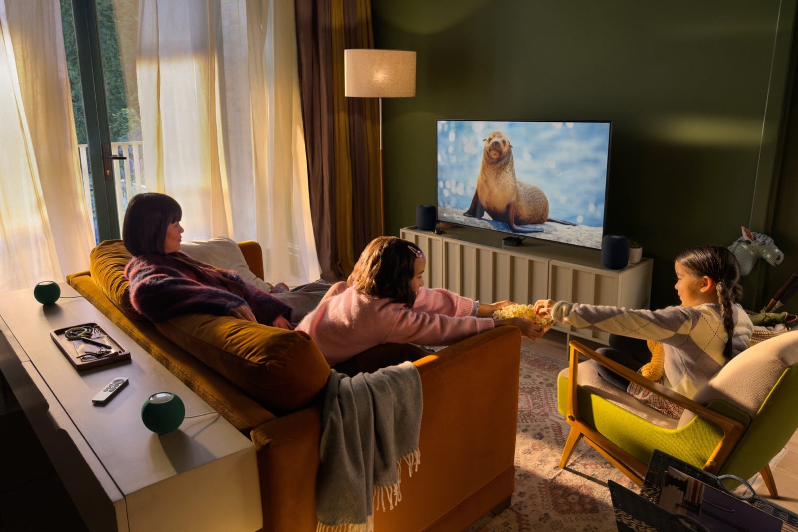 Three people watch television with two HomePods beside the screen and two smaller speakers behind the seating.