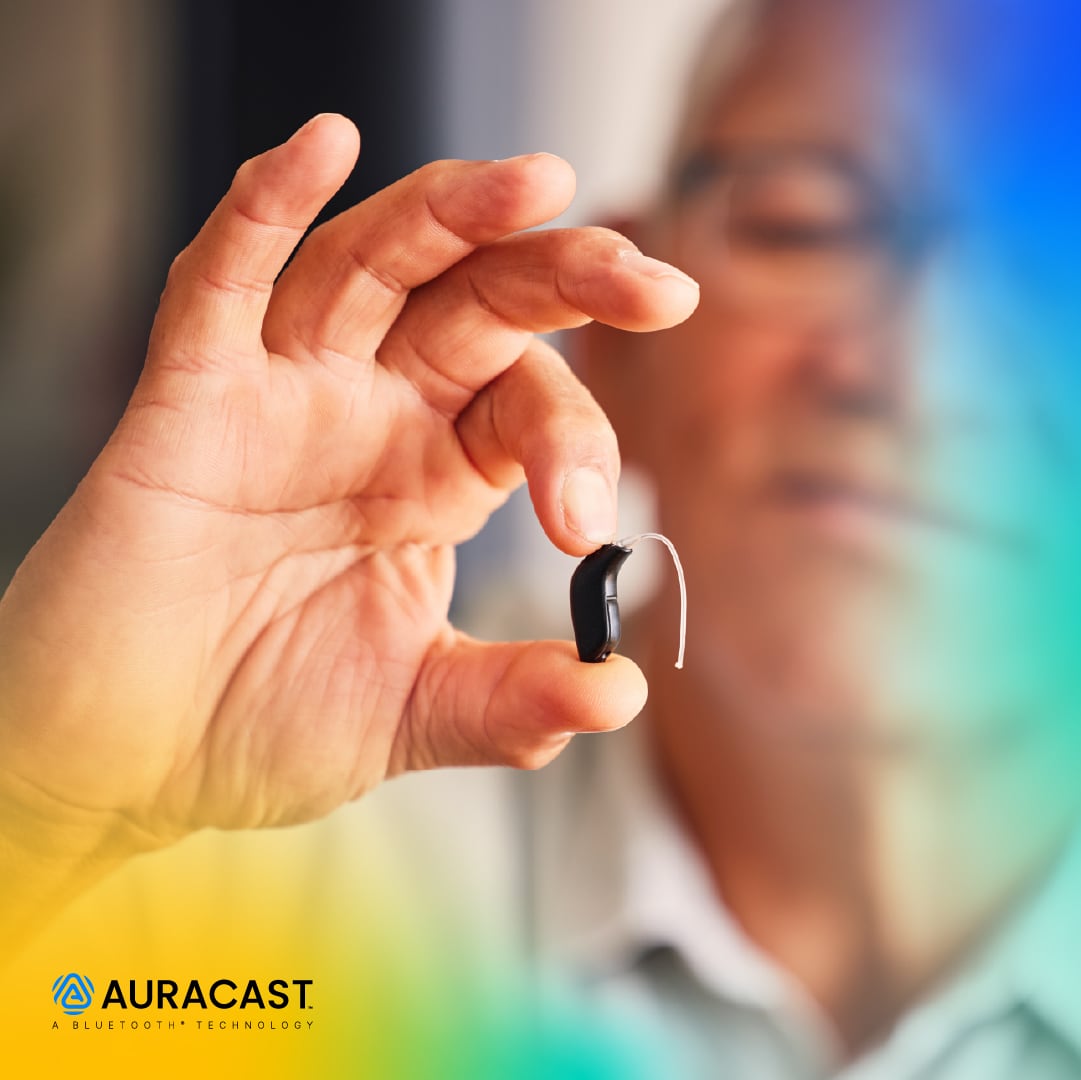 A person holds a black hearing aid between two fingers, with Auracast branding in the lower left.