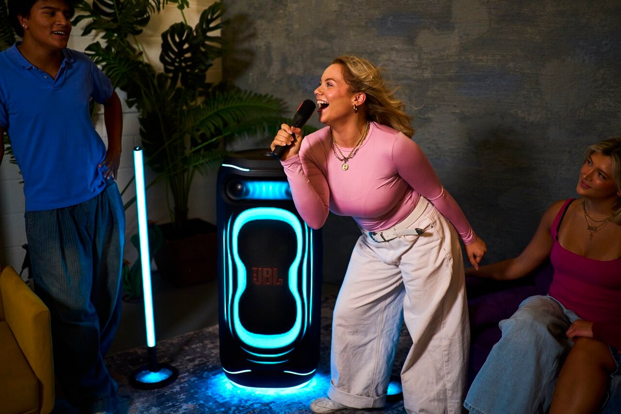 A person sings into a microphone beside a JBL speaker glowing blue and a vertical light.