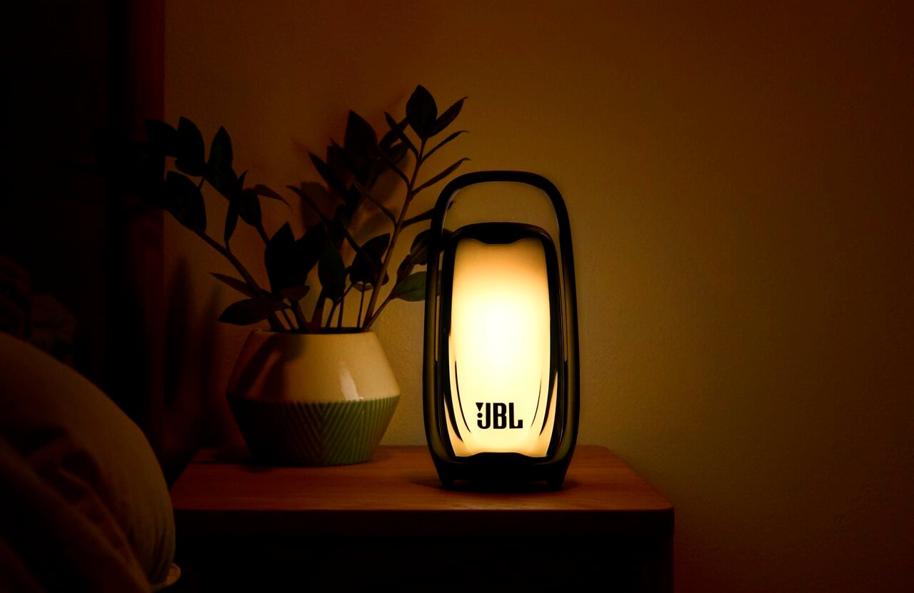 A JBL speaker glows warm yellow on a wooden bedside table beside a potted plant.