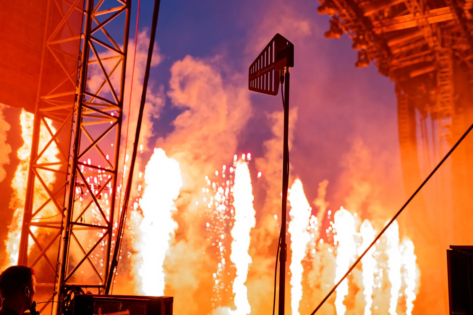 A Sennheiser antenna stands beside stage trusses as flames and sparks rise behind it.