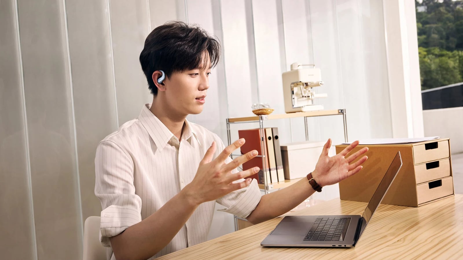A person wears a clip-style earbud while gesturing with both hands at a laptop.