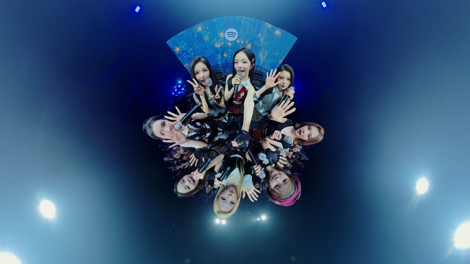 Hearts2Hearts members gather around a fisheye camera with microphones under blue stage lighting.